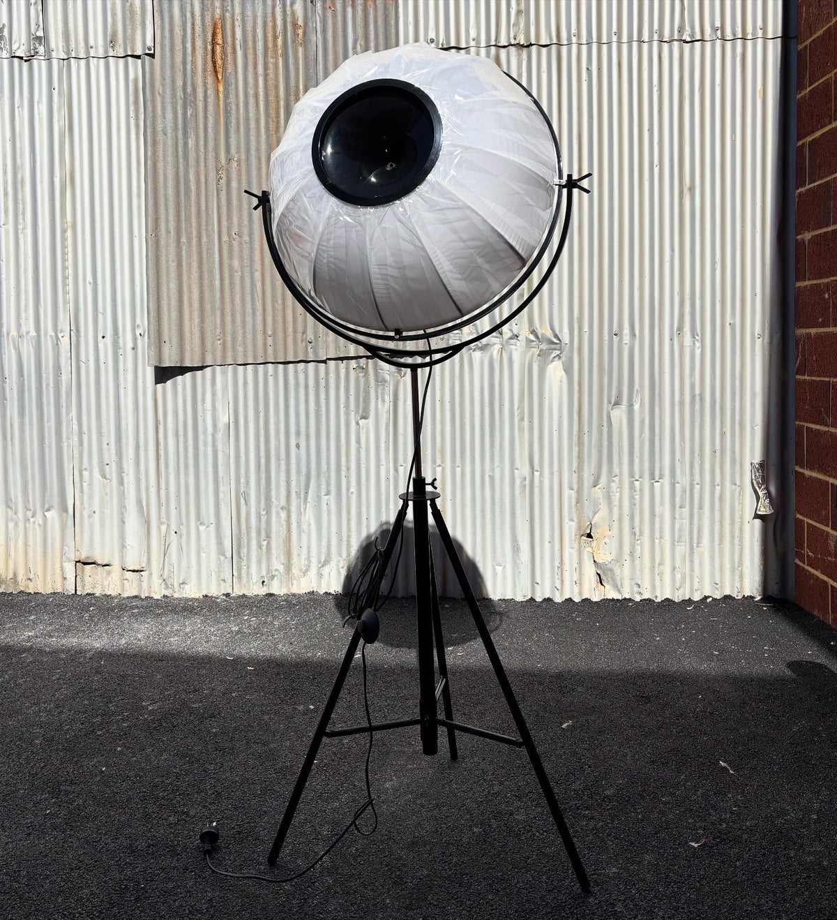 A floor lamp with a unique design featuring a black tripod base and a lampshade resembling an eye, situated against a corrugated metal background.