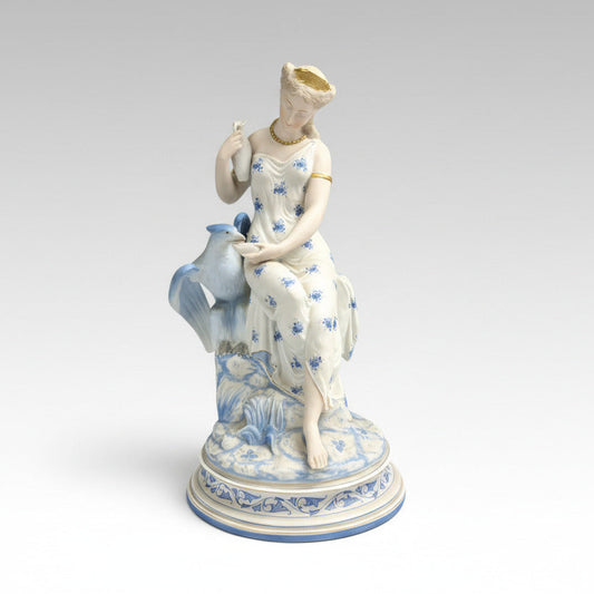 A porcelain figurine of a Neoclassical goddess with blue and gold details, seated with a bird, with a decorative blue and gold base.