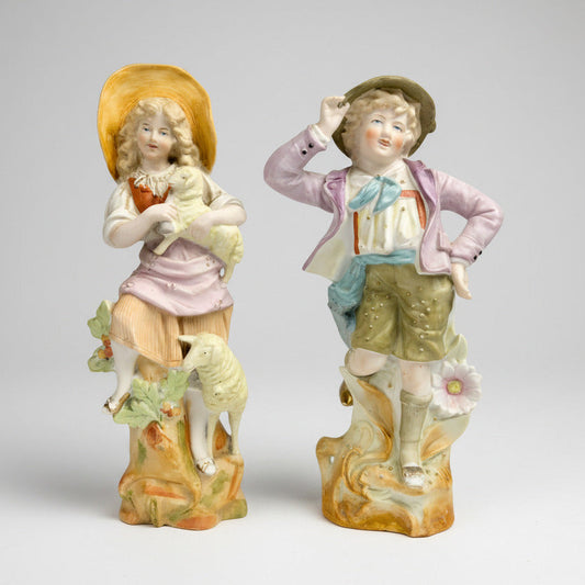A pair of German bisque figurines featuring a girl in a sun hat and a boy in a hat and shorts, with a sheep and a book.