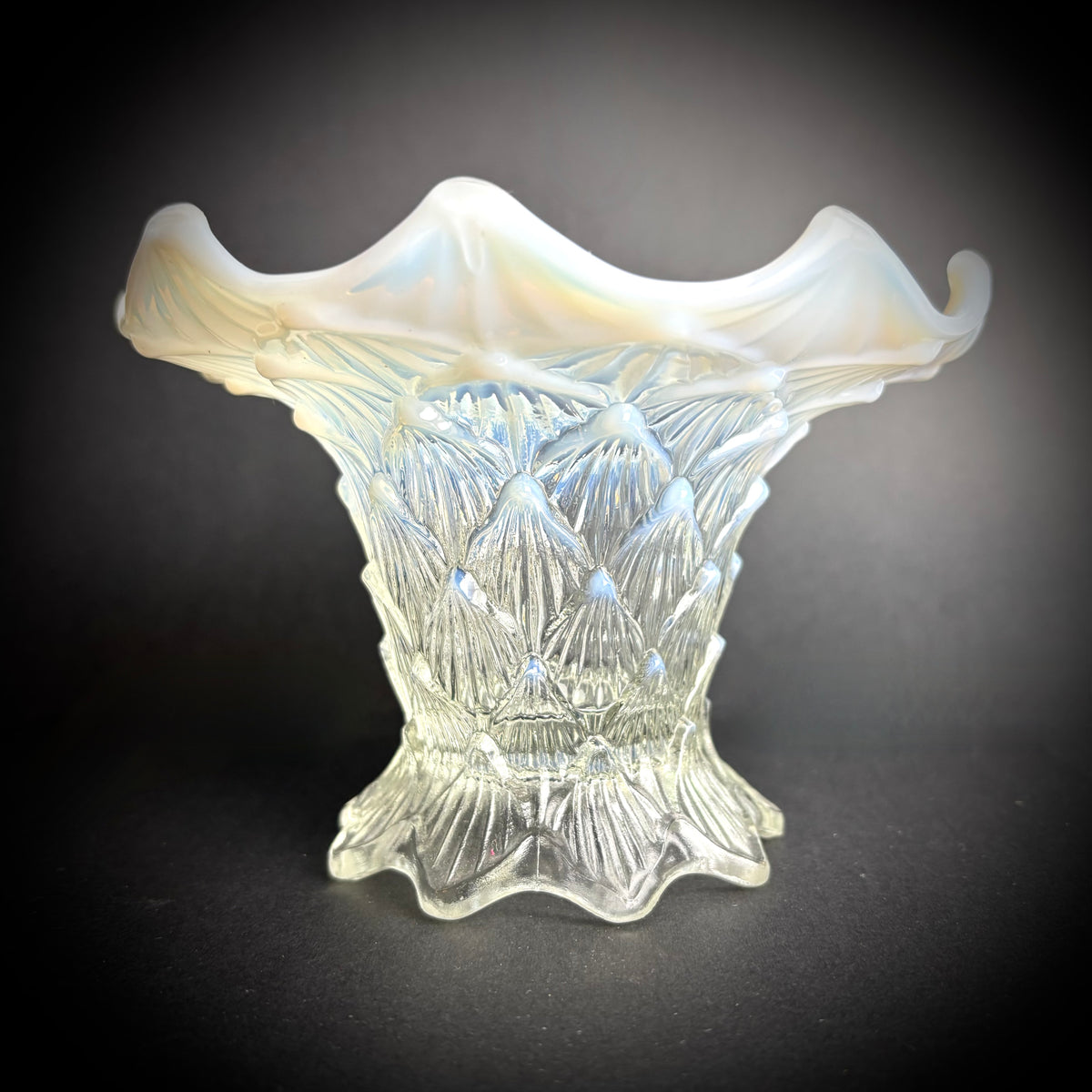 A white opalescent glass vase with a floral pattern and a scalloped edge, displayed against a dark background.