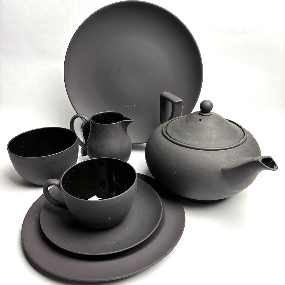 A 22-piece Wedgwood Black Basalt Tea Set displayed, including a teapot, creamer jug, sugar bowl, cups, saucers, side plates, and a cake plate, all in black color.